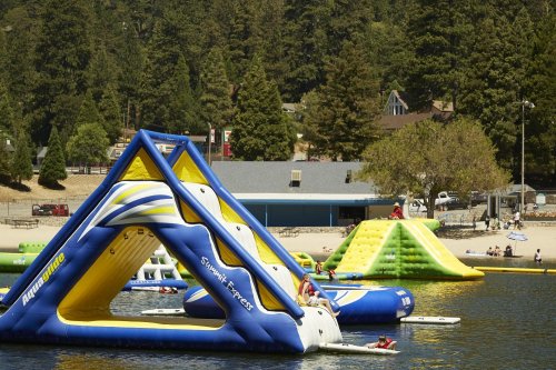 Colorful inflatable water structures on a lake, with people enjoying the beach and trees in the background.