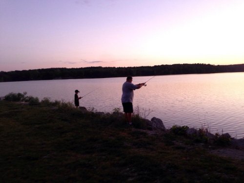 Two people fishing by a calm lake at sunset, with trees in the background and a colorful sky.