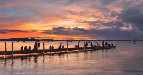 A sunset over a calm waterway, with people gathered on a wooden pier silhouetted against vibrant clouds.