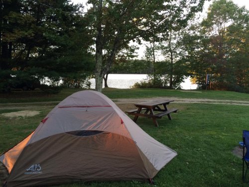 A camping scene featuring a tent on green grass, with a picnic table and a serene lake in the background.