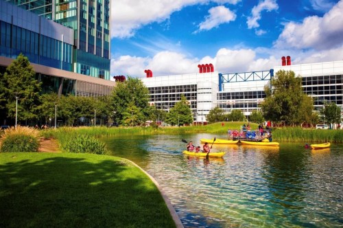 People kayaking on a serene lake surrounded by greenery and modern buildings under a blue sky with fluffy clouds.