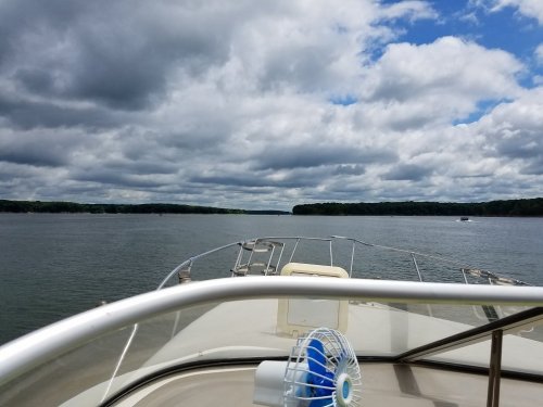 View from the bow of a boat on a calm lake, with cloudy skies and green trees lining the shore.