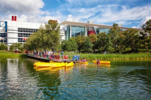 A serene lake with yellow kayaks and people paddling, surrounded by greenery and a modern building in the background.