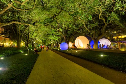 A vibrant park at night, featuring illuminated spheres and lush trees, with people walking along a pathway.
