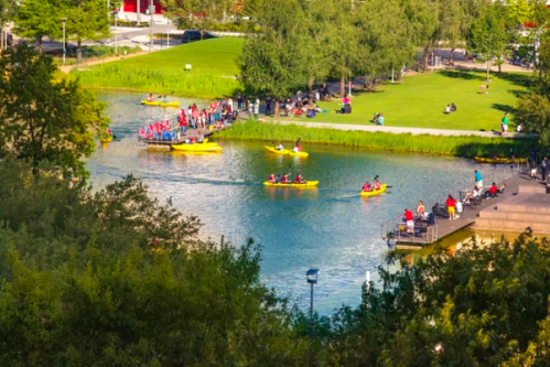 Aerial view of a park with people kayaking on a river and others relaxing on the shore. Lush greenery surrounds the area.