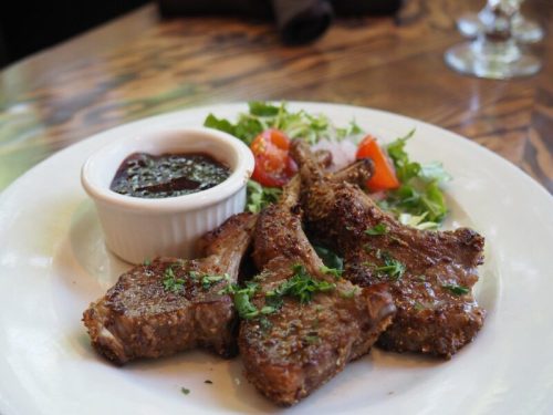 A plate of grilled lamb chops garnished with herbs, served with a side salad and a small bowl of sauce.