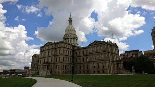 The Michigan State Capitol building with a dome, surrounded by green grass and a cloudy blue sky.