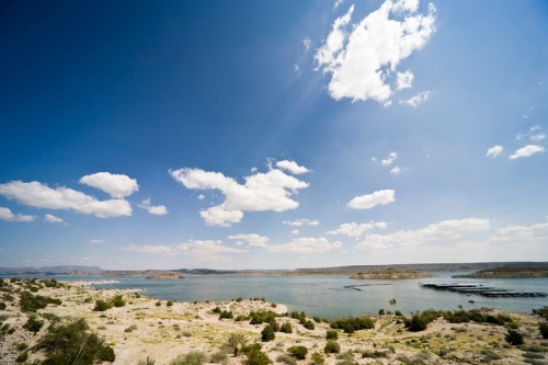 A serene landscape featuring a calm lake surrounded by rocky shores and a clear blue sky with scattered clouds.
