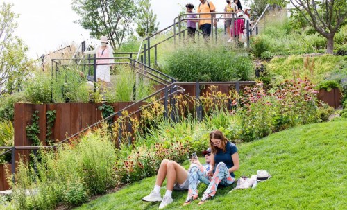 A woman sits on grass, using her phone, while others walk up a path surrounded by lush greenery and flowers.