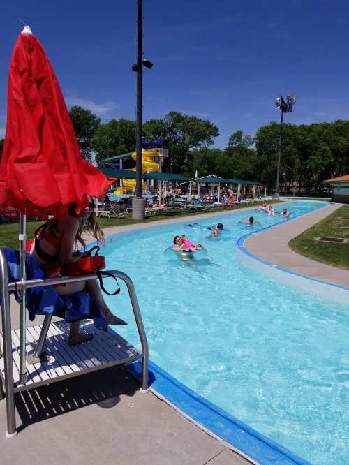 A sunny pool scene with a lazy river, water slides, and people enjoying the water under a red umbrella.