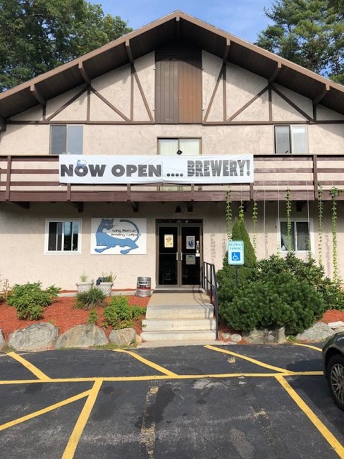 A building with a large "Now Open... Brewery!" sign, surrounded by greenery and a parking lot.