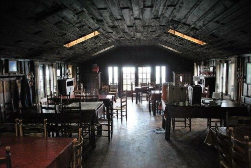 Interior of a rustic dining room with wooden tables and chairs, featuring large windows and a sloped ceiling.