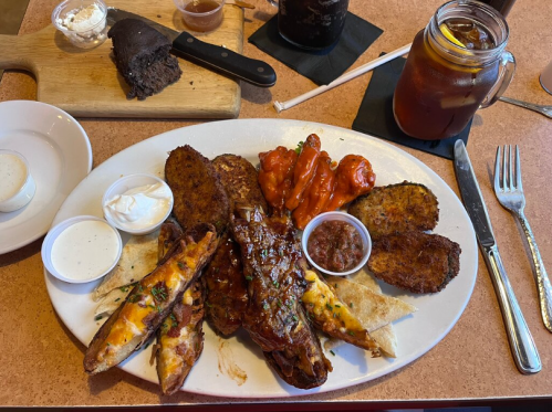 A plate of assorted fried foods, including ribs, wings, and potato wedges, with dipping sauces and a drink on the side.