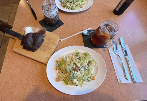 A table setting with a salad, a slice of chocolate cake, and drinks, including iced tea and a dessert in jars.