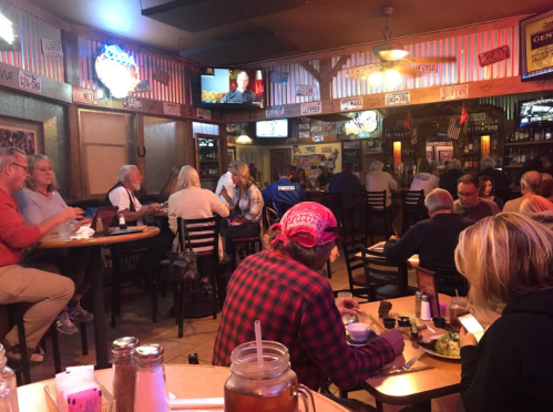 A busy restaurant scene with diners at tables, colorful decor, and a TV in the background.
