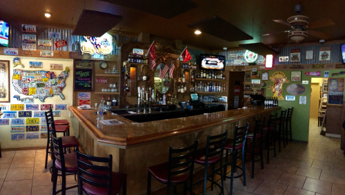 A cozy bar interior with a wooden counter, empty chairs, and colorful decor featuring signs and memorabilia on the walls.