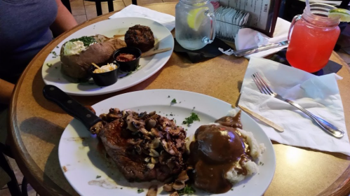Two plates of hearty meals: one with steak topped with mushrooms and mashed potatoes, the other with a baked potato and meatloaf.