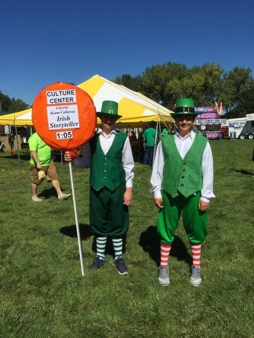 Two boys in green leprechaun costumes stand beside a sign for an Irish storyteller event at a festival.