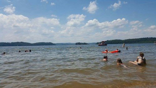 People swimming in a lake on a sunny day, with boats and hills in the background under a partly cloudy sky.