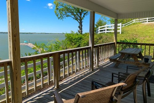 A wooden porch with seating overlooks a calm lake and green hills under a clear blue sky.