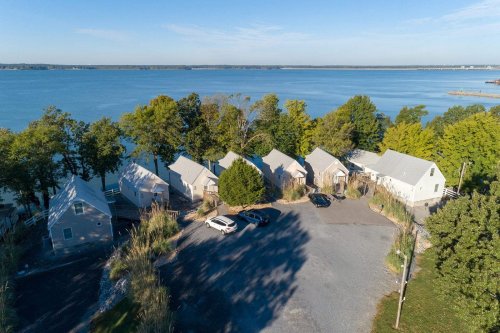 Aerial view of a cluster of cottages by a calm lake, surrounded by trees and a gravel parking area.
