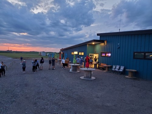 A group of people gathers outside a blue building at sunset, with clouds in the sky and a grassy field nearby.
