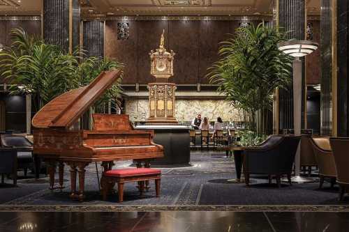 Elegant hotel lobby featuring a grand piano, ornate clock, lush plants, and a stylish bar area in the background.