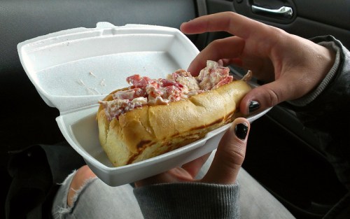 A hand holding a lobster roll in a takeout container, with a car interior in the background.