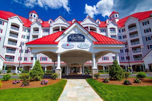 A large, white hotel with a red roof and multiple stories, surrounded by greenery and flowers under a blue sky.