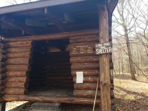 A rustic log cabin shelter with a sign reading "Long Pond Trail Shelter" in a wooded area.