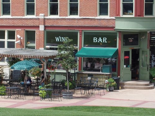 Outdoor seating area of a wine bar with green umbrellas, surrounded by brick buildings and potted plants.