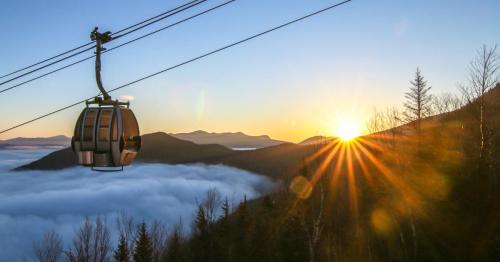 A gondola glides above a sea of clouds at sunrise, with mountains silhouetted in the background.