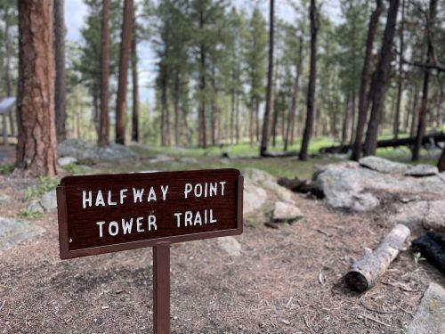 A wooden sign reading "Halfway Point Tower Trail" in a forested area with trees and rocks in the background.