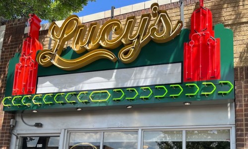 Sign for "Lucy's" with bright yellow lettering and red decorative elements, set against a green background.