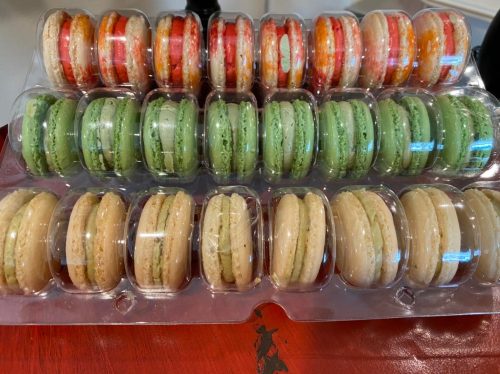 A tray of colorful macarons in various flavors, arranged in rows with red, green, and beige hues.