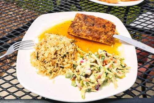A plate with seasoned fish, rice with herbs, and a colorful coleslaw on a black metal table.