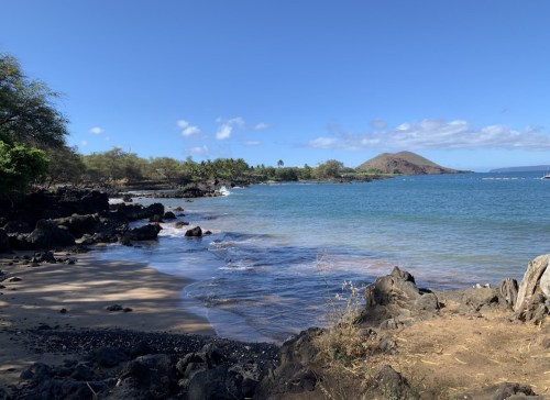 A serene beach scene with rocky shores, clear blue water, and a distant hill under a bright blue sky.