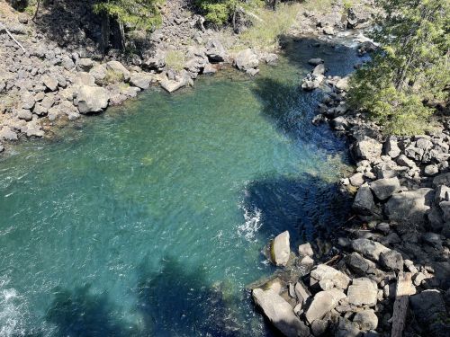 A serene river pool surrounded by rocks and trees, with clear blue water reflecting sunlight.