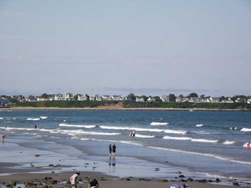 A beach scene with people walking along the shore, gentle waves, and houses lining the distant coastline.