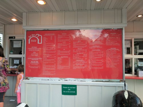 A red menu board displaying food options at a takeout stand, with a child and adult waiting nearby.