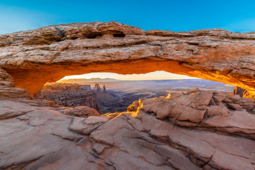 A stunning rock arch frames a panoramic view of a desert landscape at sunset, highlighting rugged cliffs and distant formations.