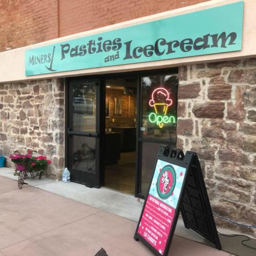 Exterior of Miners Pasties and Ice Cream shop, featuring an "Open" neon sign and a colorful A-frame menu board.