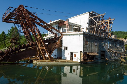 A large, rusted dredge boat sits on calm water, surrounded by trees and a clear blue sky.