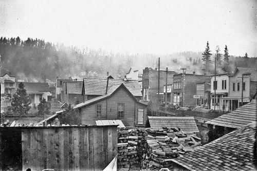 Historic black-and-white photo of a small town with wooden buildings and a forested backdrop, shrouded in mist.