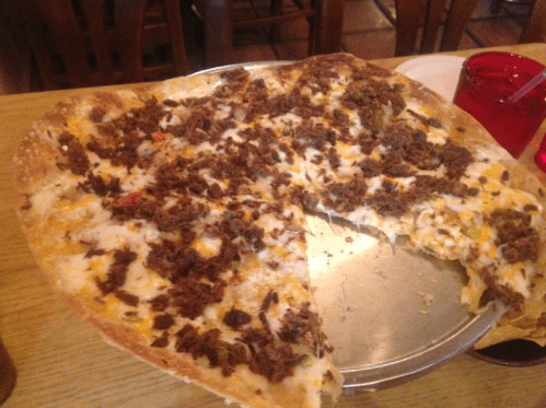 A large pizza with a thick crust topped with cheese and ground meat, partially sliced on a metal tray.