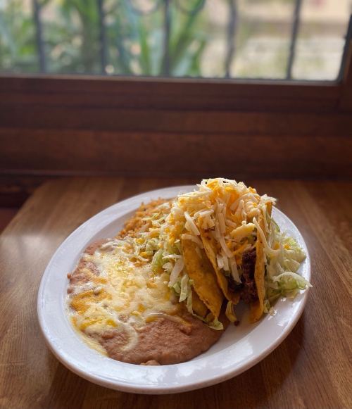 A plate of tacos with shredded lettuce and cheese, served with refried beans and a side of rice, near a window.
