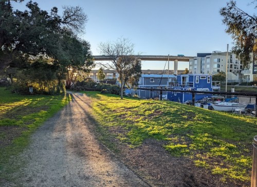 A sunlit path through a park, with greenery and a view of buildings and boats in the background.