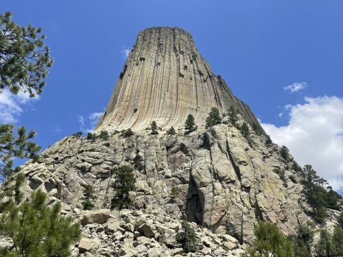 A towering rock formation rises against a blue sky, surrounded by trees and rocky terrain.