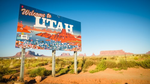 A colorful "Welcome to Utah" sign with desert landscape and rock formations in the background under a clear blue sky.