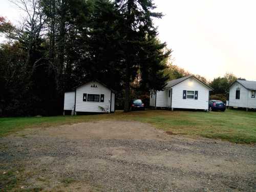 Two small white cabins surrounded by trees, with a gravel area in front and parked cars nearby.